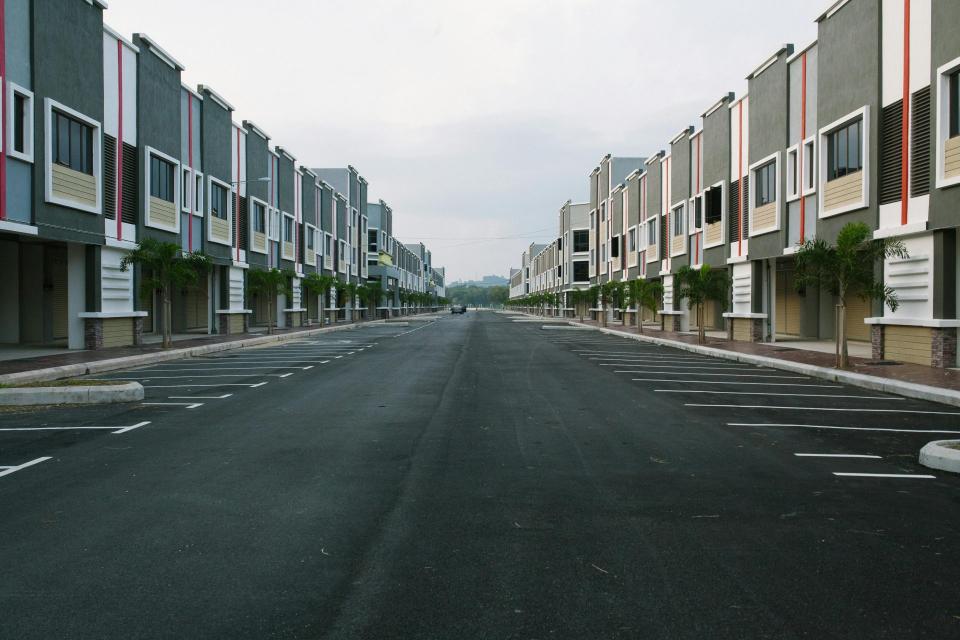 A quiet, empty urban street flanked by modern condominiums under a cloudy sky, with ample parking space.